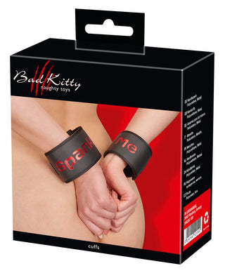 En låda Badkitty Handbojor - Spank Me, med svarta manschetter med djärv röd "Spank Me"-text, är elegant uppvisad på en modells handleder, vilket ger en vågad touch till alla BDSM-lekar-kollektioner.