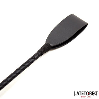 A Svart BDSM Piska 60cm PU Läder med texturerat handtag ligger på vitt underlag och utstrålar dominans; Latetobeg BDSM-line logotyp syns i hörnet.