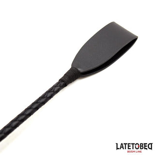 A Svart BDSM Piska 60cm PU Läder med texturerat handtag ligger på vitt underlag och utstrålar dominans; Latetobeg BDSM-line logotyp syns i hörnet.