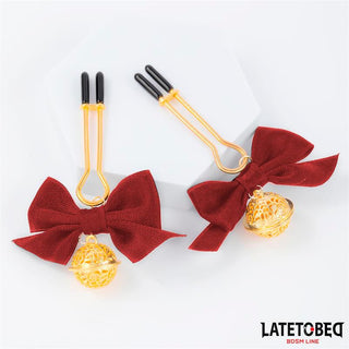 Nippelklämmor med röd rosett: Två guldklämmor med justerbara spetsar för individuell komfort, prydda med röda rosetter och guldfiligranklockor. Utformade för BDSM-lek. Visas på en vit bakgrund med LATETOBEG-logotypen.