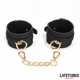 Handbojor Med Guldfärgad Kedja: Ett par svarta texturerade bondage-manschetter med guldringar och en guldkedjekoppling, perfekt som en snygg bondage-tillbehör, visas på en vit bakgrund.