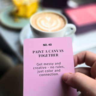 En hand håller ett Undressed Intimacy Cards Kortlek-kort med texten "Paint a canvas together" framför en latte, perfekt för att stärka relationen.