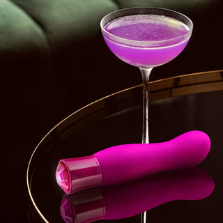 En lila cocktail i ett glas ligger bredvid den eleganta rosa Exclusive Tourmaline Vibrator på ett runt, reflekterande bord.