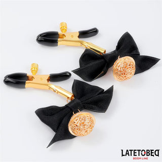 Nipple Clamps With Bow har justerbara klämmor i guld med svarta rosetter och dekorativa gulddetaljer - perfekta för BDSM-lek och elegant presenterade på en vit bakgrund.