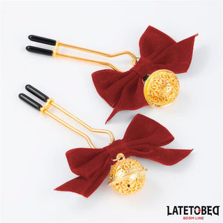 Red Bow Nipple Clamps har en guldfärgad metallkonstruktion, röda rosetter och eleganta guldfiligranberlocker. De justerbara klämmorna erbjuder en anpassningsbar passform och tillför stil och funktion till din BDSM-samling på en vit bakgrund.