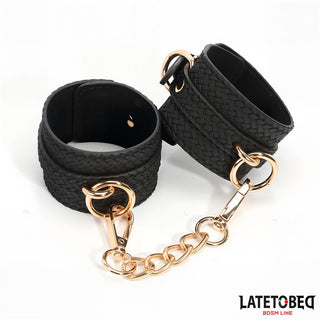 Handbojor Med Guldfärgad Kedja: Svarta vävda handledsbojor förbundna med guldkedja och spänne - en elegant bondageaccessoar. LATETOBED BDSM LINE-logga i hörnet.