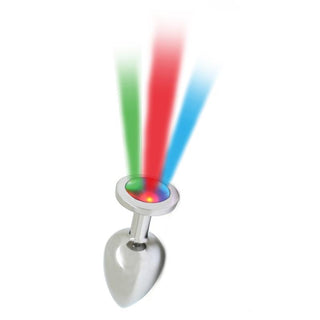 Rimba Buttplug Med LED, ett känsligt silver hjärtformat metallobjekt med en slät yta, avger livfulla röda, gröna och blå ljusstrålar från toppen och kastar ett förtrollande sken mot den enkla vita bakgrunden.
