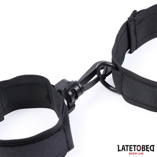 Svart justerbart nylonhalsband med handledsfästen med metallklämmor och Latetobeg BDSM Lines logotyp i hörnet.