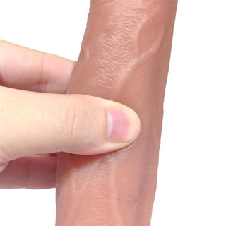 En hand klämmer ihop ett texturerat, cylindriskt föremål som liknar flytande silikon. Det ljusbruna föremålet efterliknar Realistisk Turbo Dildo med Rörelse, med venliknande mönster för flexibilitet. En tumme och ett pekfinger trycker försiktigt för att framhäva dess realistiska design.