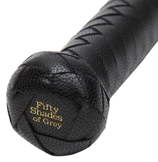 En närbild av Fifty Shades of Grey Flogger avslöjar dess svarta läderpiskahandtag med korssömmar, med "Fifty Shades of Grey" präglade i guld i slutet, som förkroppsligar sensuella dominans.
