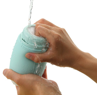 Händerna håller försiktigt och tvättar en Tenga Puffy Masturbator Mint Grön, som visar upp sin mjuka silikon-design, under rinnande vatten mot en orörd vit bakgrund.