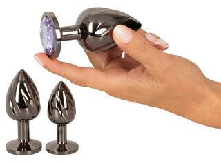 En hand håller ett glänsande föremål från Metall Butt Plug Set, med en lila pärla på änden. I närheten sitter två liknande metallpluggar upprätt, något mindre och idealiska för temperaturspel eller analsöjning.