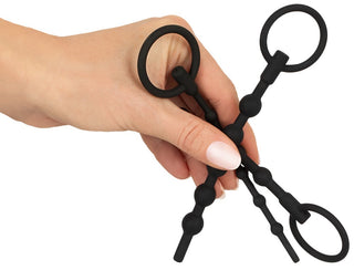 En hand håller två föremål från Urethral Dilator Set 3-pack, med svarta silikonbitar med cirkulära ringar i varje ände, som påminner om ridverktyg. Deras design inkluderar en serie rundade pärlor längs deras längd.
