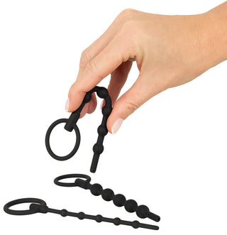 En hand håller svarta analpärlor av silikon med ett ringhandtag, med två liknande set på en vit yta. I närheten kompletterar Urethral Dilator Set 3-pack arrayen och erbjuder olika alternativ för entusiaster.