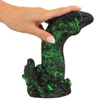 En hand håller i Beasty Cocks Lizard Peak, en reptilinspirerad dildo gjord av flytande silikon i grönt och svart. Den har en strukturerad, tentakelliknande design med böljande mönster och en virvlande bas.