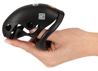 En hand håller i Cock Cage med Dilator - en svart, böjd bur med hål, en synlig QR-kod på ytan och plats för en silikon-dilator.