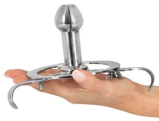 En hand håller i Anal Spreader Deluxe Stål, en mekanisk anordning av metall med en cylindrisk mittdel och krokförsedda stöd.