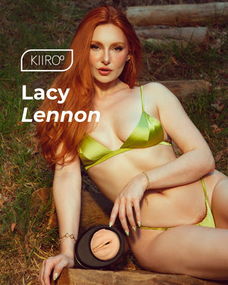 En kvinna med långt rött hår i gröna underkläder poserar utomhus och håller i en rund behållare för Feel Lacy Lennon Masturbator.