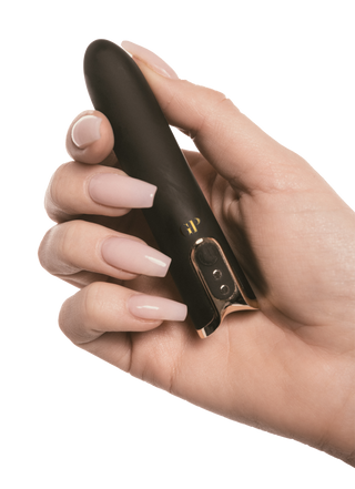 En hand med manikyrerade naglar håller Crave Bulletvibrator, en elegant USB-laddningsbar enhet i svart och guld, mot en vit bakgrund.