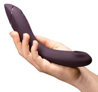 En person håller Womanizer OG, en böjd lila enhet med knappar, designad för dubbelstimulans och med olika vibrationslägen, mot en vit bakgrund.