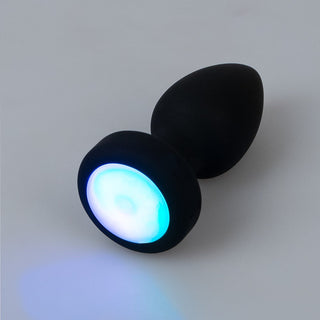 LED Buttplug med Vibration i svart silikon med lysande blå och vit LED-lampa vid basen, visas mot en vanlig ljus bakgrund.