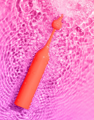 ROMP Pop klitorisvibrator i orange silikon med vattendroppar, med en böjd design som visas på en rosa krusad yta.