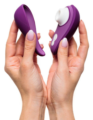 Två händer visar den lila Womanizer Liberty 2 Luftvibratorn och framhäver dess avtagbara silikondelar. Den har Pleasure Air Technology med en vit cirkulär sektion och knappar på ena halvan, med en elegant, böjd design.
