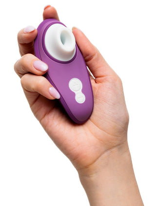 En välvårdad hand håller den lila Womanizer Liberty 2 Luftvibrator, designad med Pleasure Air Technology. Med en rund vit öppning och två centrala vita knappar står den elegant mot en orörd vit bakgrund.