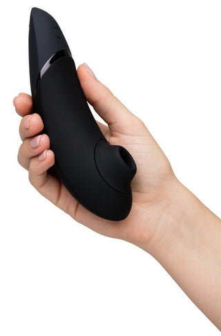 En hand håller Womanizer Next 3D Luftrycksvibrator, en elegant svart enhet med en böjd design, knapp och rund spetsöppning, med avancerad Smart Silence-teknologi. Bakgrunden är vanlig vit.