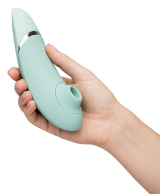 En hand håller Womanizer Next 3D Luftrycksvibrator, en mintgrön, böjd enhet med Smart Silence-teknologi, med en cirkulär öppning i ena änden och en metallisk accent nära toppen.