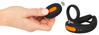 En hand håller en svart och orange fjärrkontroll bredvid den eleganta Vibrerande Scrotumstimulator Rebel, vilket tyder på en förbättrad upplevelse.