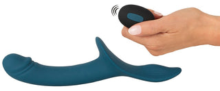 En hand håller en liten svart fjärrkontroll bredvid en blå, ergonomisk vibrator med ett böjt skaft och strukturerad bas, en del av Strap-On Set med Vibrator. Fjärrkontrollen visar upp sin trådlösa funktionalitet.