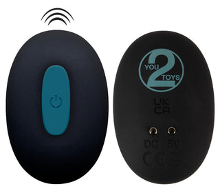 Två svarta ovala elektroniska enheter visas. Den vänstra påminner om en fjärrkontroll med blå strömknapp och signallinjer. Till höger visas märket "Strap-On Set med Vibrator", certifieringsmärken och laddningsportikoner.