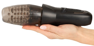 En hand håller Stroker Penis Masturbator, en svart cylindrisk enhet med en transparent texturerad ände utformad för roterande och stötande funktioner.