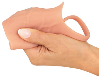 En hand klämmer ihop den beige Penis Sleeve Öppen Realistixxx, ett silikonliknande texturerat föremål med ett öglehandtag. Den flexibla och töjbara designen påminner om en penishylsa, vilket gör den spännande.