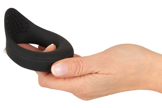 En hand håller i Vibrerande Ballring Rebel, en sexleksak i svart silikon som är formad som en ring med texturerade ojämnheter på ena sidan.