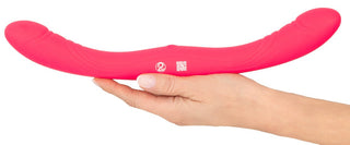 En hand håller i Couples Choice Double Vibrator, en lång, rosa silikonleksak med dubbla ändar, mot en vit bakgrund.