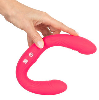 En hand håller i Couples Choice Double Vibrator, en flexibel rosa silikonleksak, mot en vit bakgrund.