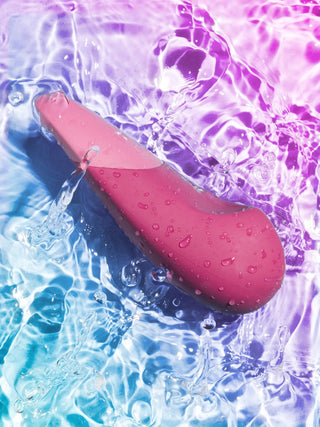 Ett rosa föremål med vattendroppar är nedsänkt i porlande lila och blått vatten, som liknar en Womanizer Vibe Klitorisvibrator som visar upp sin UltraWave-teknologi.