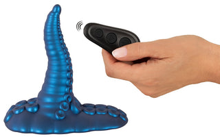 En hand håller i en Tentacle Pad Sit On Vibrator med sugkoppar, formad som en blå tentakel och utrustad med en fjärrkontroll.