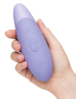 En hand håller en lila, ovalformad Womanizer Enhance Luftvibrator, med kraft-, öknings- och minskningsknappar. Enheten har en slät finish med rundade kanter och innehåller Pleasure Air Technology för en förbättrad användarupplevelse.