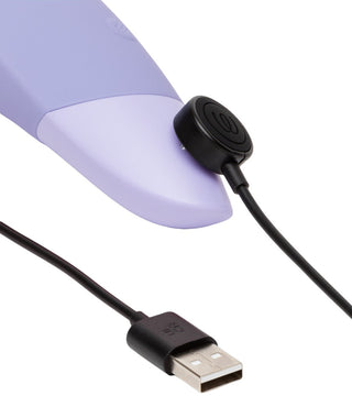 Närbild av den lila Womanizer Enhance Luftvibrator med Pleasure Air Technology, ansluten till en USB-laddningskabel. Kabeln har ett magnetiskt cirkulärt fäste som ansluter till enheten, med USB-kontakten synlig i andra änden.