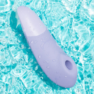 Womanizer Enhance Luftvibrator, en ljuslila och vattentät enhet, stoltserar med en elegant design som flyter i klart blått vatten. Droppar glittrar på dess yta, vilket framhäver dess vattentäta kvalitet. Förbättrad med Pleasure Air Technology, garanterar den en oöverträffad upplevelse.