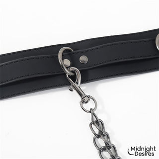 Svart halsband med tillhörande fotbojor i veganskt läder, sammanlänkade med en metallkedja och prydda med logotyp.