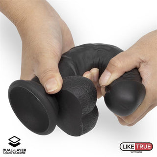 Händerna sträcker en svart, flexibel LikeTrue Realistisk Dildo 20 cm med en sugbas, vilket visar dess flytande silikonkvalitet, medan logotyper i hörnen framhäver varumärket med två lager silikon.