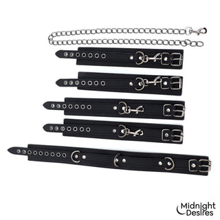Svarta handledsbojor i veganskt läder med spännen, metallringar och en kopplingskedja, avbildade mot en vit bakgrund.