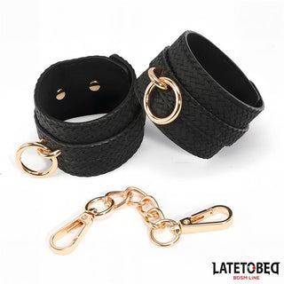 Handbojor Med Guldfärgad Kedja: Två svarta texturerade bondage-manschetter med guldringar och en matchande guldkedja, ett slående BDSM-tillbehör på en vit bakgrund.