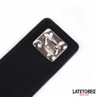 En närbild av Svart Kyskhetsbälte Kvinna Justerbart, med ett svart PU-läderband med en D-ring i metall och LATETOBEG BDSM LINE-logotypen - perfekt för att ge en djärv BDSM-stil till alla kollektioner.