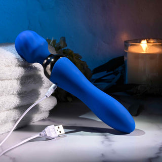 En Selopa Blue Belle Wand, USB-uppladdningsbar, vilar på en marmoryta bredvid vita vikta handdukar och ett tänt ljus, vilket skapar en lugn och avkopplande atmosfär.