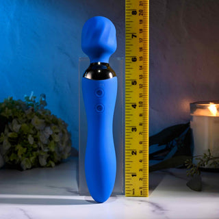 En Selopa Blue Belle Wand med dubbelsidig design står upprätt bredvid ett gult måttband som avslöjar att den är cirka tio tum hög. Den mjukt upplysta scenen inkluderar grönska och ett tänt ljus, vilket gör denna USB-laddningsbar massageapparat perfekt för att lägga till lugn i alla rum.